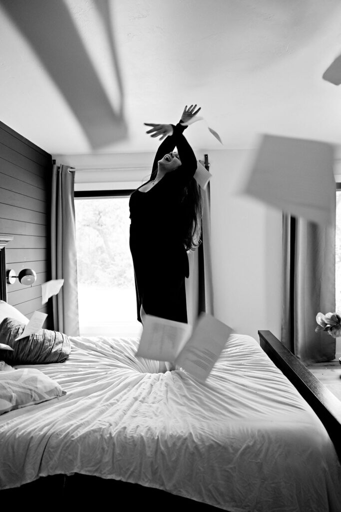Mom dancing joyfully on bed with pages of poetry flying through the air. A mother dances freely on a bed while pages scatter around her — representing the joy and liberation of analog self care and creative expression. Used in blog post: "Your Phone Break Isn't Actually a Break — A Self Care Reset for Working Moms."
