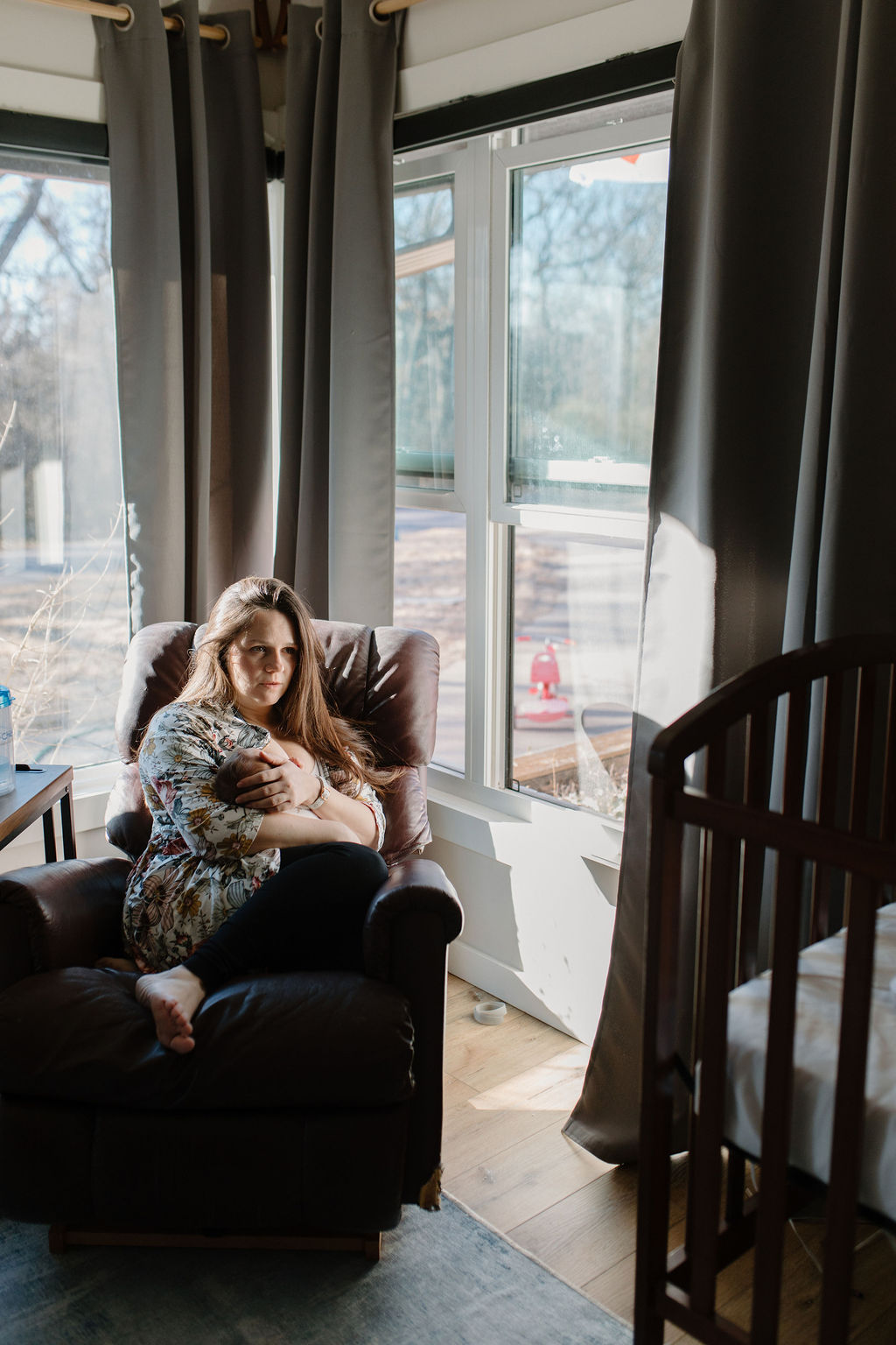 Working mom in floral robe nursing infant, exhausted and staring into the distance